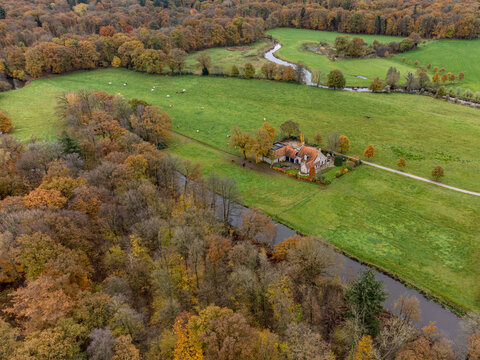 Aerial view of a charming house nestled amidst the vibrant hues of autumn foliage, bordered by lush green fields and a meandering river, Tilligte, Netherlands.