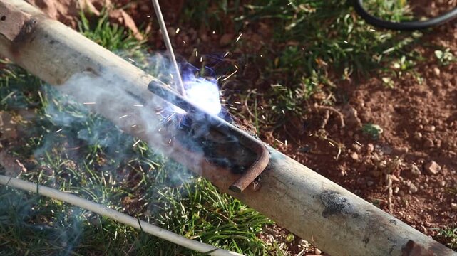 A welder is performing arc welding on an iron pipe in an open area.