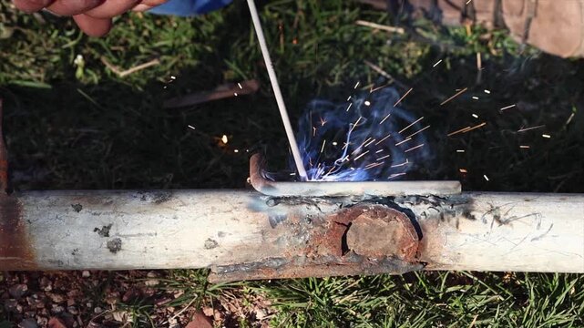 A welder is performing arc welding on an iron pipe in an open area.