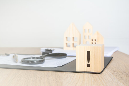Single exclamation mark on a wooden block. Blurred background with miniature houses, magnifying glass and contract. Real estate warning concept.