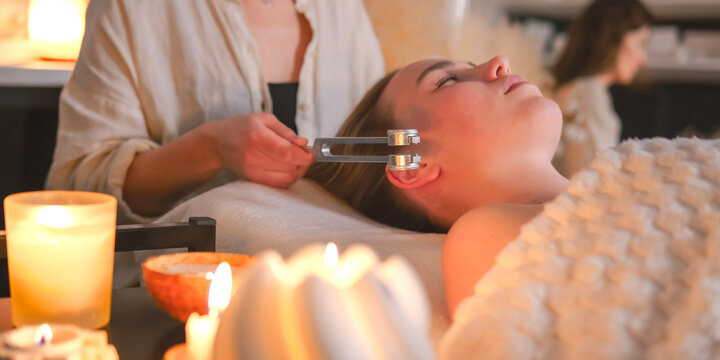 Tuning fork sound therapy with candles, practitioner applying vibration to woman's neck, holistic wellness ritual, relaxation, alternative spa treatment, close up.