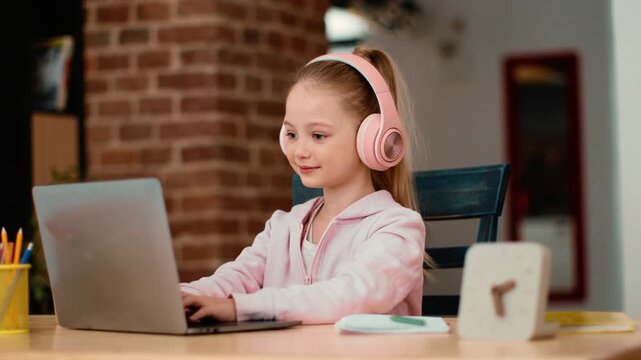 A young girl with light skin is sitting at a desk, focused on her laptop. She wears pink headphones and types intently while surrounded by school supplies in a cozy study space.