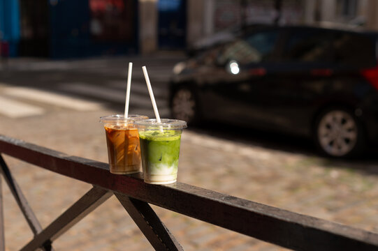 Iced matcha latte and iced coffee in plastic cups with white straws on a black metal railing. Street background with cobblestones and a blurred car in Paris, France.