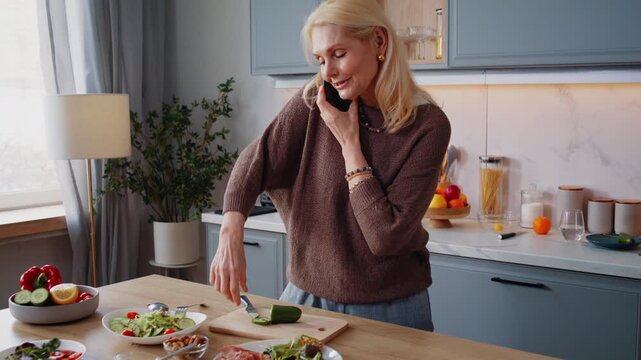 Cheerful elderly woman answers phone call as stands near counter