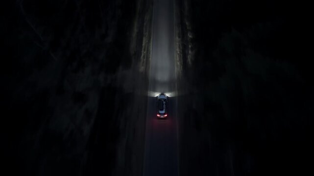 Aerial night shot of a car driving through dark forest with headlights on as drone follows vehicle along straight road