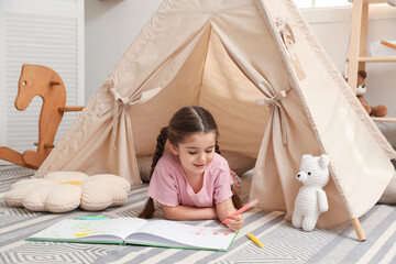 Little girl drawing in sketchbook near toy wigwam at home © New Africa