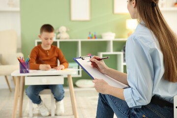 Professional psychologist taking notes while little boy drawing at white table indoors, selective focus