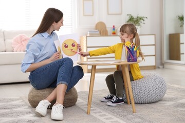 Professional psychologist showing pictures of sad and happy faces to little girl at wooden table indoors