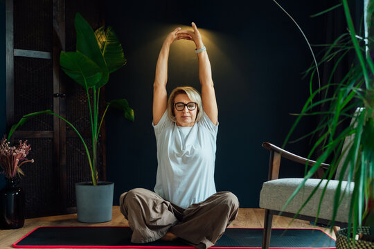 Middle aged woman on floor mat and doing stretching yoga position in modern Scandi dark interior home. Healthy Lifestyle. Sport activity for mature wellbeing. Mindful movement and relaxation