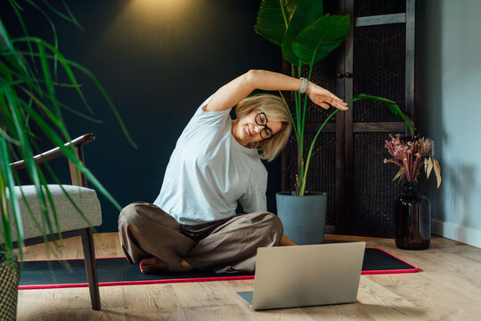 Middle aged woman practicing online yoga workout using laptop in modern Scandi dark interior home. Healthy Lifestyle. Sport activity for mature wellbeing. Fitness online training. Mindful movement