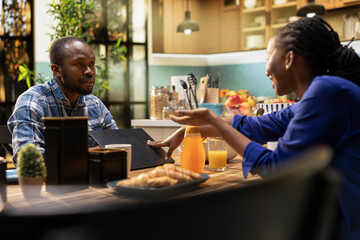 Fototapeta premium African american woman asking her boyfriend to stop working at breakfast, needing some attention while they drink coffee and serve food at home. Busy man always doing remote work.