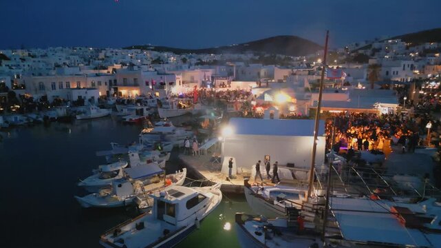 Evening aerial drone view of Naousa village on Paros island, Greece, featuring illuminated streets, coastal harbor and festive atmosphere during a local celebration. Ideal for travel, culture
