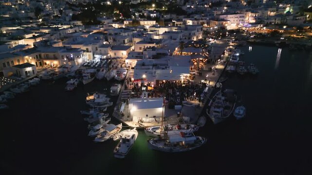 Evening aerial drone view of Naousa village on Paros island, Greece, featuring illuminated streets, coastal harbor and festive atmosphere during a local celebration. Ideal for travel, culture