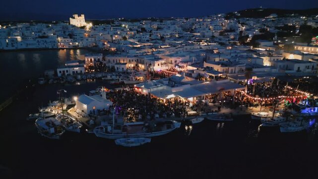 Evening aerial drone view of Naousa village on Paros island, Greece, featuring illuminated streets, coastal harbor and festive atmosphere during a local celebration. Ideal for travel, culture