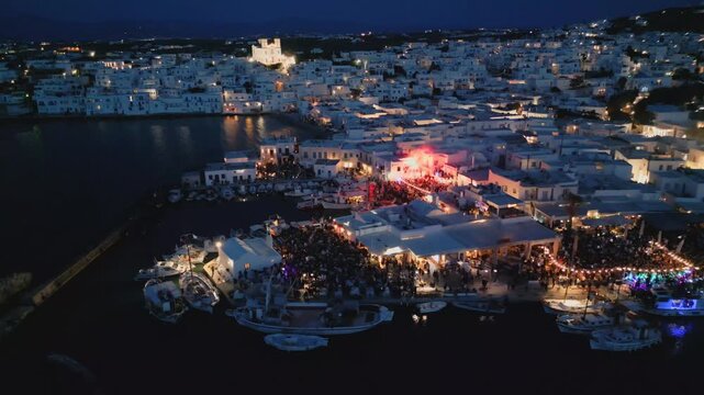 Evening aerial drone view of Naousa village on Paros island, Greece, featuring illuminated streets, coastal harbor and festive atmosphere during a local celebration. Ideal for travel, culture