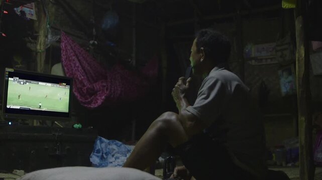 Man in a modest home in Myanmar watching a live soccer match on television at night
