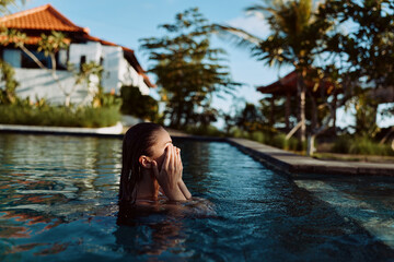 Fototapeta premium Person submerged in a pool, hair wet, hands shielding eyes, beside a resort villa under a blue sky. Lifestyle scene focused on relaxation, leisure, and warm water.
