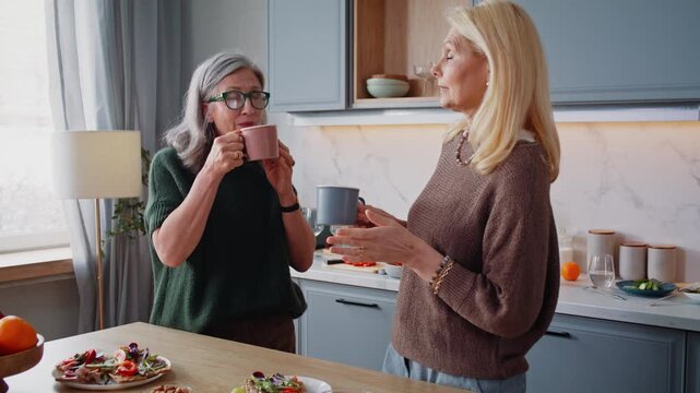 Mature women friends with cups talk sharing news in kitchen