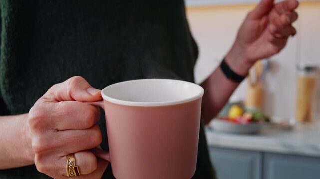 Senior woman hand holds cup of hot tea as stands in kitchen