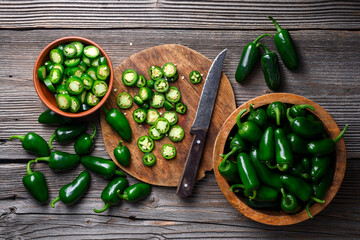 Jalapeno peppers with bowl of slices and knife on wooden surface. Fresh green chili cooking scene...