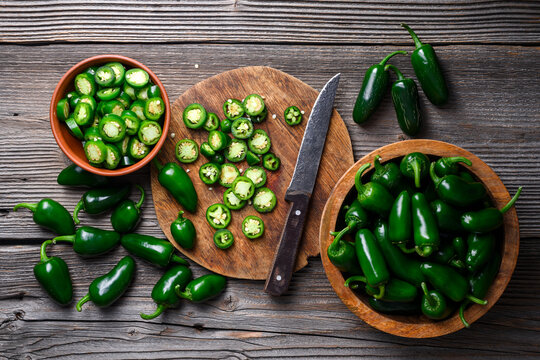 Jalapeno peppers with bowl of slices and knife on wooden surface. Fresh green chili cooking scene rustic composition