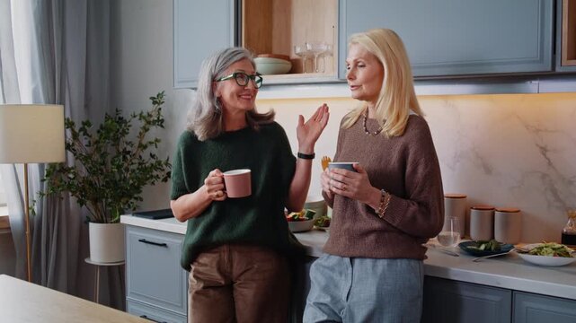 Cheerful mature women with tea cups talk standing in kitchen