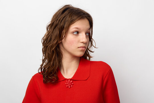Young woman with curly brown hair wearing a red sweater looks to the side with a neutral expression on a plain white background. Concept of casual style and natural beauty.