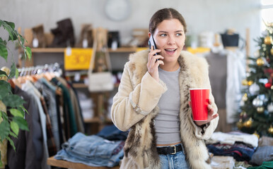 Fototapeta premium European girl with cup of coffee talking on phone in womens clothing store. Shopping as hobby and pastime. Megamall, sales and promotions, offline store of branded clothing manufacturer