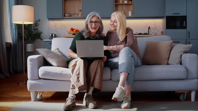 Senior women smile at laptop together while sitting on sofa