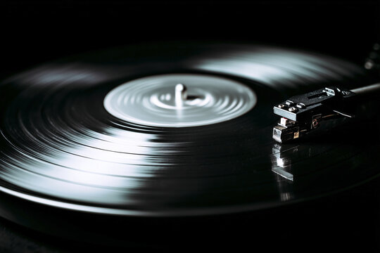 Close up of a spinning black vinyl record on a turntable with a needle headshell. Atmospheric dark background with elegant reflections, representing retro music and audiophile culture