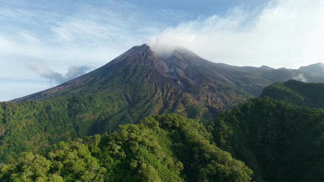 An aerial view of Mount Merapi surrounded by dense forest in Java, Indonesia. A view of volcanic terrain and hills under a clear blue sky.
