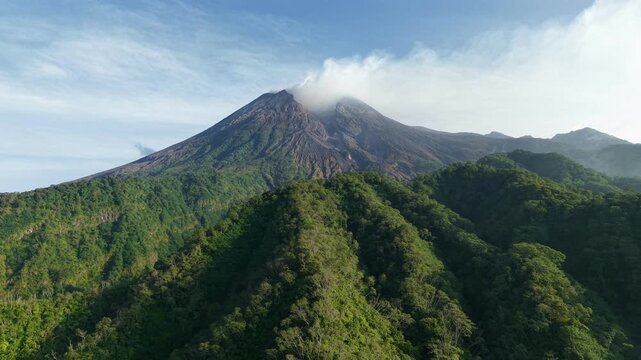 Drone footage shows Mount Merapi spewing smoke surrounded by dense forest in Java, Indonesia. The volcanic landscape and hills appear mysterious under a clear blue sky.