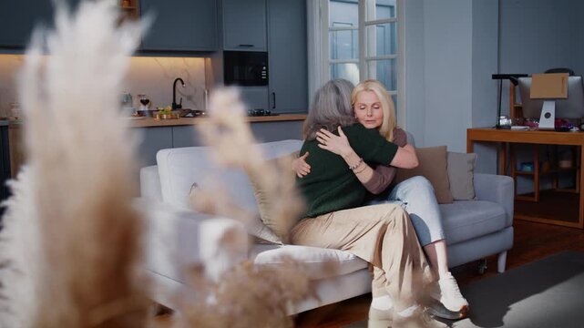 Mature women hug warmly on cosy sofa during reunion at home