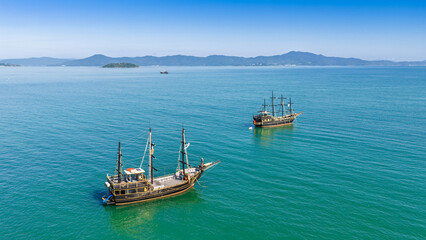 Two tall-ship pirate vessels anchored on the turquoise waters of Florianopolis bay under clear blue sky. © Marcio Eneas
