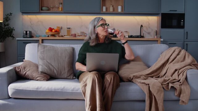 Elderly woman smiles drinking wine and holding laptop on sofa