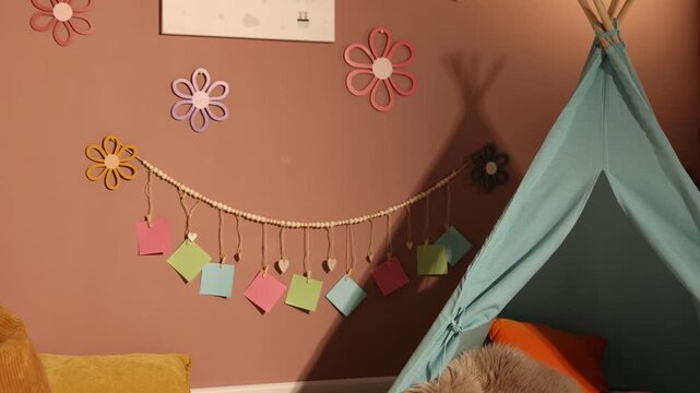 Colorful paper notes and beautiful decor hanging on brown wall near toy wigwam in stylish children's room, camera moving forward. Interior design