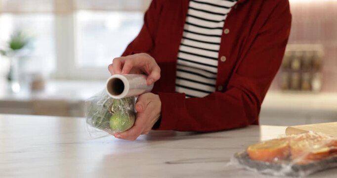 Woman wrapping fresh cucumbers in cling film at light marble table, closeup