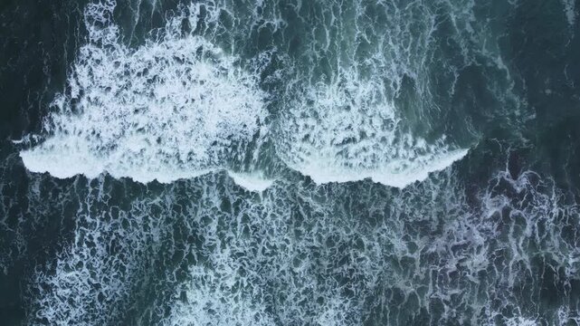 Aerial view of waves crashing against the jagged black lava shoreline of the Indian Ocean coast, creating dramatic white foam patterns in the deep blue waters.
