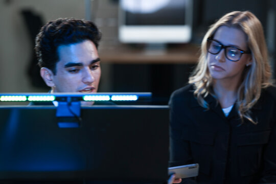 Dynamic tech team collaborates late night. Young Asian woman points at screen explaining project details to male colleague reviewing documents in modern hub workspace.