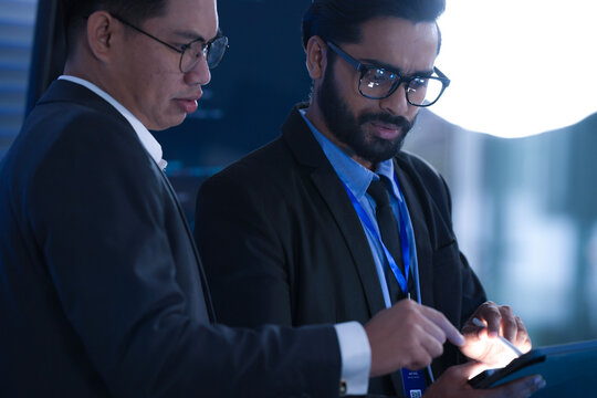 Dynamic tech team collaborates late night. Young Asian woman points at screen explaining project details to male colleague reviewing documents in modern hub workspace.
