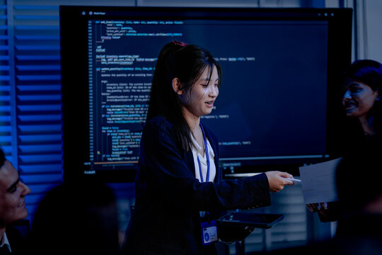 An Indian female developer leads a code review, holding a report. She explains a Python script for an AI or cybersecurity system to her team in a dark SOC Security Operations Center