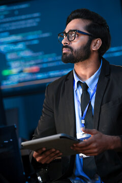An Indian male software engineer or data scientist holds a tablet in a command center. He is asking a question about the AI code or cybersecurity data shown on the large screen.
