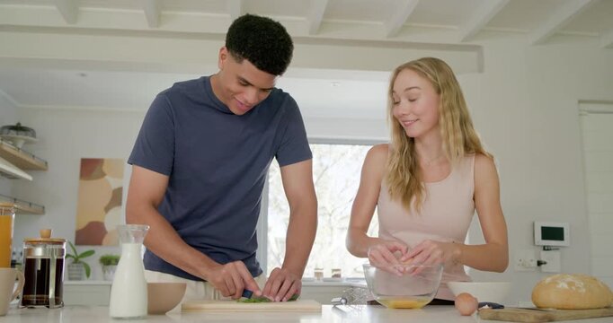 Diverse couple making breakfast at kitchen island, chopping herbs on board, cracking eggs in bowl