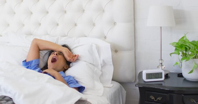 Indian woman waking up after digital alarm ringing, yawning and rubbing eyes in bed