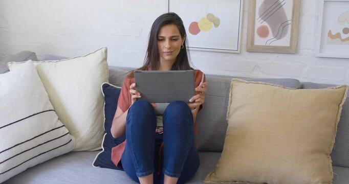 Indian woman sitting on gray sofa holding tablet in folio shifting tablet checking phone for alert
