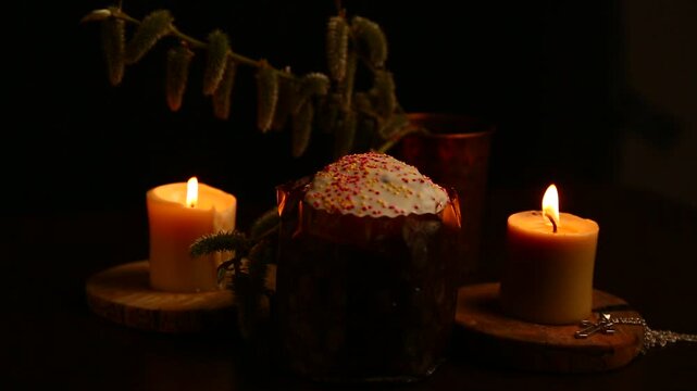 Two bright Easter burning candles with Easter cake on dark black background on natural wooden candlesticks, silver holy cross, willow branch on a table. Closeup warm concept, Easter holiday. No people