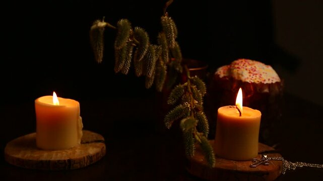 Two bright Easter burning candles with Easter cake on dark black background on natural wooden candlesticks, silver holy cross, willow branch on a table. Closeup warm concept, Easter holiday. No people