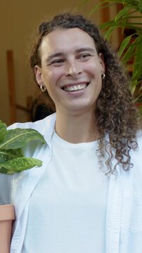 Vertical video: Holding plant, person in white tee laughing at offscreen voice in entryway with pot