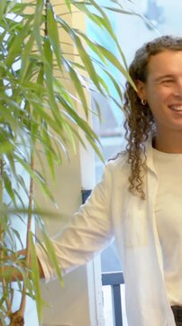 Vertical video: Entering right, adult calling friend on phone at door touching plant in white tee