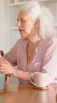 Vertical video: Leaning senior in pink top examining watch to help relative offering wrist at home